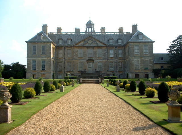 Image of a beautiful Belton House in Lincolnshire, an avenue with topiary bushes either side, leading up to a grand four-story building with wings to the right and left, and a central entrance up a shallow set of steps.