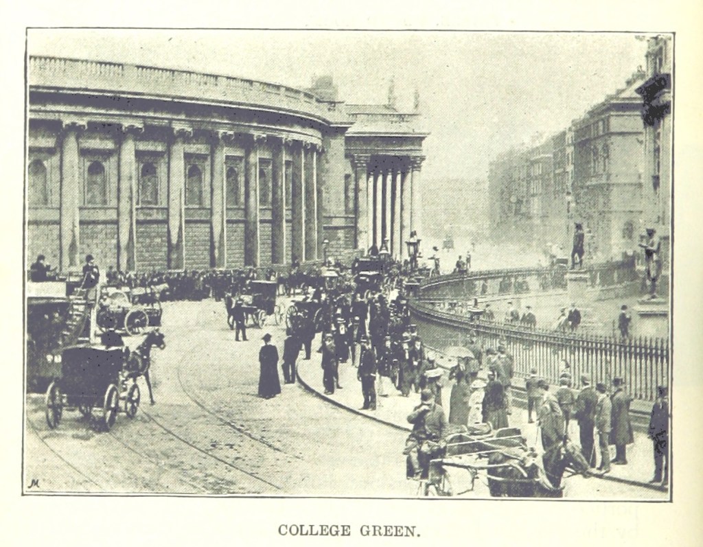 View of a Dublin street, containing a large number of pedestrians and several carriages. The gates of Trinity College are visible to the right and the Bank of Ireland Building is at the front and centre of the image.