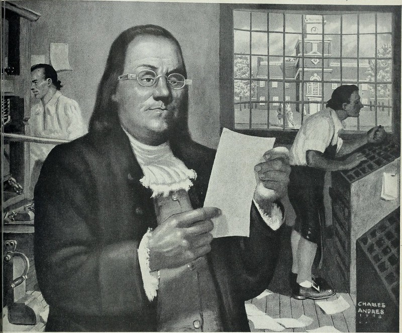 A reproduction of a famous portrait of Benjamin Franklin, represented at a printing office. Two men are working in the background: one is setting type and the other is handling a press. Franklin stands in the foreground, dressed in a coat and ruffled neckerchief and wearing glasses. He is holding up a piece of paper and his expression is confused, which is probably an unintentional result of the picture being reproduced for a magazine.