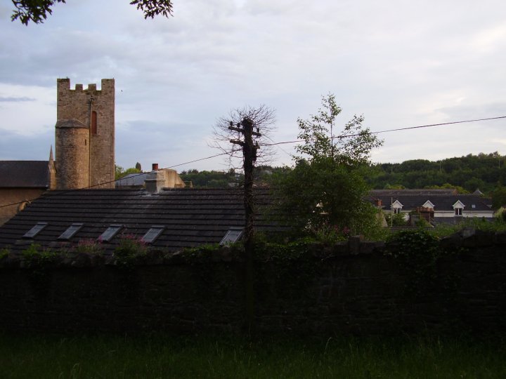 View towards Chapelizod from the park. An old stone wall, part of the park boundary, is in the foreground, with grass in front. Behind it can be seen the roofs of modern houses and an electrical pole. To the left of the image rises the medieval tower of St. Laurence, and other parts of the church building. In the distance can be seen more trees, on the far side of the Liffey.