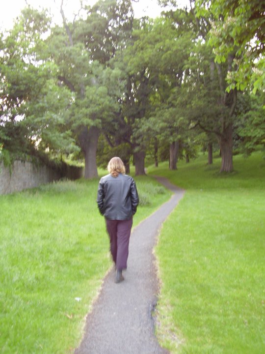 A man in a short black jacket walks down a gravel path bordered on either side by grass. He is facing away from the camera, and trees can be seen further down the path. A grey stone wall is visible to the left of the image, overgrown by moss and plants.
