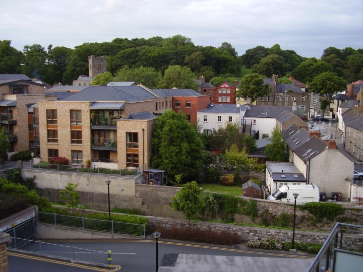 The village of Chapelizod, seen from a four-floor balcony. In the foreground is a narrow branch of the river Liffey, with modern houses and apartment buildings just beyond. Behind these can just be seen a row of buildings on Chapelizod Main Street, and the medieval church tower with crenellations and narrow windows. Above these are a hill and the treeline of the Phoenix Park.