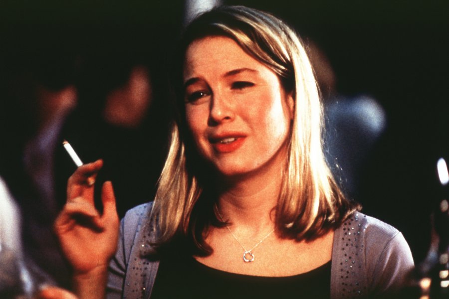 The image shows Bridget Jones standing in a black t-shirt and grey cardigan, holding a cigarette in her left hand. In the background there are some blurry shadows of people. By Nicholas R. Andrew. Source: https://openverse.org/image/9a058285-1ec9-4f6d-823c-c96460b1c4ef?q=Bridget+jones&p=113 